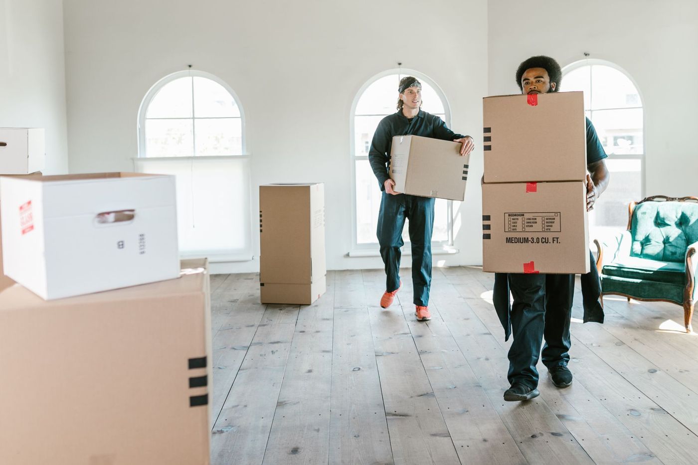 Two removal crew members carrying packed boxes through a bright, emptied living room