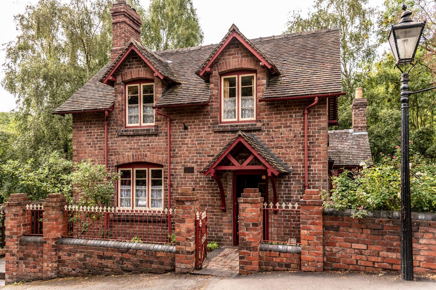 A Victorian red-brick house with gabled windows behind a low brick wall and iron gate